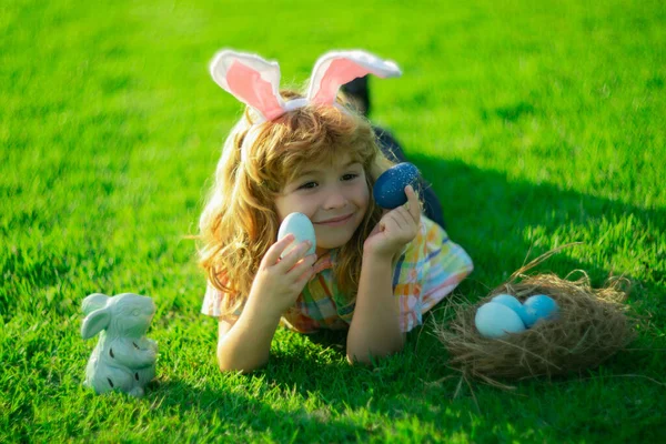 Kind met konijnenoren, paasvakantie. Hij jaagt op paaseieren. Schattig konijntje liggend op gras in park, grappig kids portret. — Stockfoto