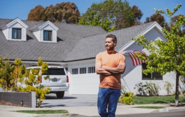 Yeni evin önünde duran yakışıklı adamın portresi. Kot pantolonlu ve tişörtlü mutlu adam Amerikan evine karşı el ele tutuşuyor..