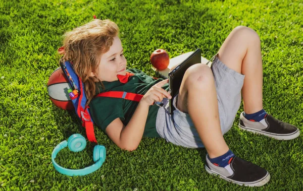 Dziecko z dodatkami szkolnymi i tabletem i odpoczywające na trawie po szkole. — Zdjęcie stockowe