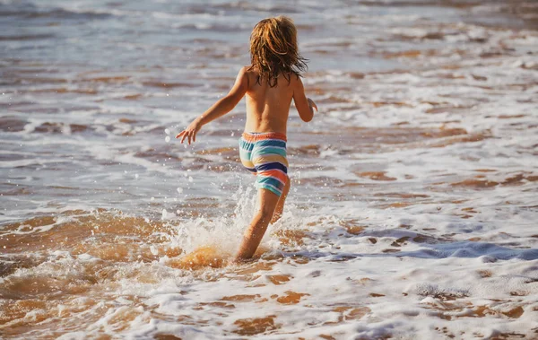 Vista trasera del niño diviértete en la playa tropical del mar. Divertido niño correr con salpicaduras por la piscina de agua a lo largo de borde de surf. Actividad infantil en vacaciones de verano. — Foto de Stock