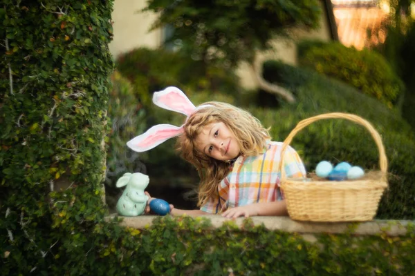 Schattig kindkonijntje draagt konijnenoren in de tuin, fijne paasdag. Konijnenjongen met paasmand, grappig buitenportret. Jachteieren en voorjaarsvakantie met kinderen. — Stockfoto