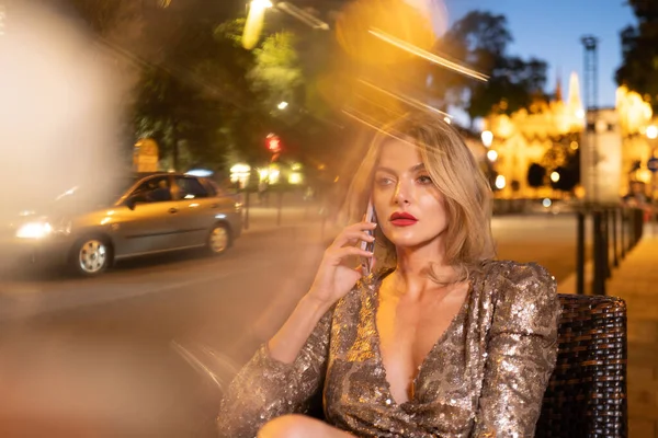 Hermosa mujer joven relajarse en la cafetería nocturna en la calle de la ciudad. Hermosa joven mujer de moda hablando en el teléfono móvil. —  Fotos de Stock