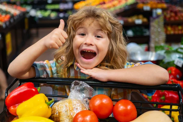 Prodej, konzumace a dítě. Nadšený kluk s jídlem v nákupním košíku v obchodě. Vtipný chlapec s palcem nahoru je nakupování v supermarketu. — Stock fotografie