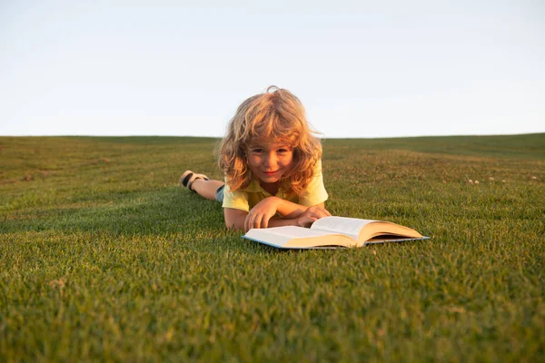 Niño inteligente leyendo libro tendido en la hierba sobre hierba y el fondo del cielo. — Foto de Stock