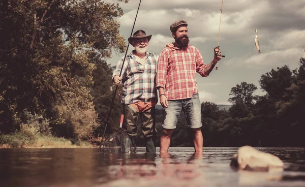 Gelukkige vader en zoon samen vissen in de zomerdag onder een prachtige hemel aan de rivier. Vliegvissen. Vliegvisser met behulp van vliegvishengel in prachtige rivier. — Stockfoto