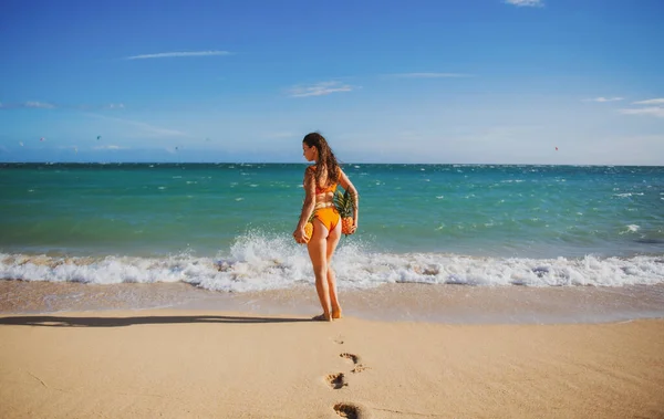 Beautiful caucasian sunbathed woman model in swimsuit posing on summer beach with white sand on blue sky on virginia or tenerife ocean background. Buttocks, butt, ass with pineapples. — Photo