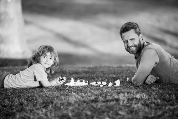 Отец и сын играют в шахматы, лежа на траве в газонном парке. День отца, любовь к семье, родительство, детская концепция. Умный ребенок во время игры в шахматы. — стоковое фото