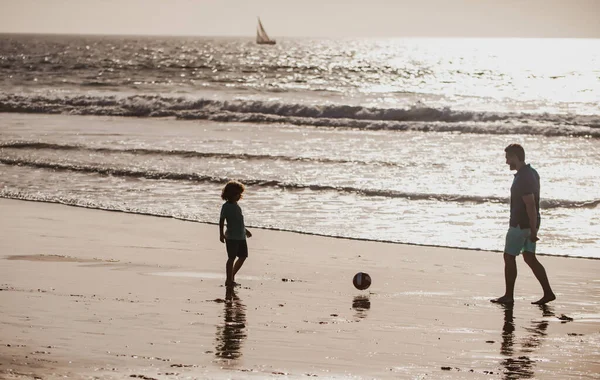 父と息子はビーチでサッカーやサッカーをします。家族旅行、休暇、父の日の概念。屋外で遊んでいる父と子供. — ストック写真