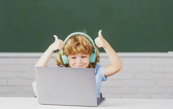 Crianças da escola com polegares para cima usando computador portátil e fones de ouvido na sala de aula na escola. — Fotografia de Stock