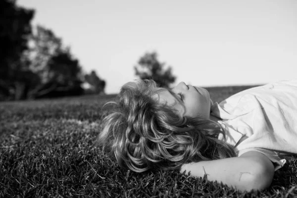 公園で草の上に寝そべっている幸せな小さな金髪の子供。学校の休暇中の夏の日。素晴らしい子供の夢と笑顔. — ストック写真