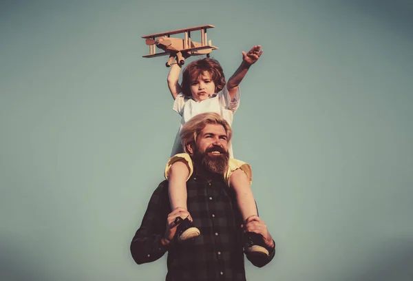 Vaderdag. Vader en zoon. Vader geeft zoon een lift terug. Portret van een gelukkige vader die zoon meelift op zijn schouders en omhoog kijkt. Leuke jongen met papa spelen met houten vliegtuig outdoor. — Stockfoto