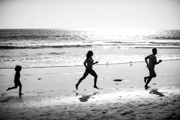 お父さん、お母さん、お子さんが海岸を走っています。健康的な家庭生活をコンセプトとする。夏の人が走り、親と子供が海でスポーティな休暇を過ごす。子供と一緒に屋外でジョギングする家族. — ストック写真