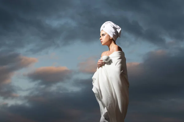 Расслабленная женщина в халате с полотенцем на голове, нюхающая кофе в ночное время на драматическом небе под открытым небом. Утренний курорт. — стоковое фото