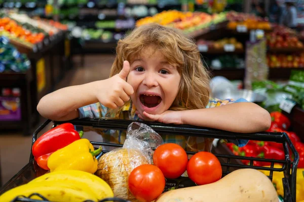 Sale, consumerism and child. Excited little kid with food in shopping cart at grocery store. Little child choosing fresh vegetables in a food store.