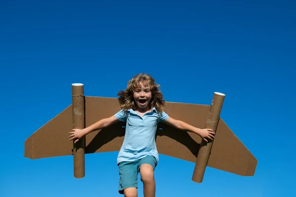 青い空に紙の翼を持つ幸せな子供。春の緑の野外で楽しいおもちゃのジェットパックを持つ子供。自由気楽で子供の想像力夢のコンセプト. — ストック写真