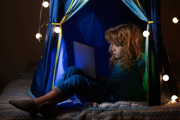 En liten pojke barn använder en bärbar dator på natten, ett barn sitter hemma tält framför en datorskärm i rummet med neon blixtnedslag, begreppet gadget missbruk. — Stockfoto