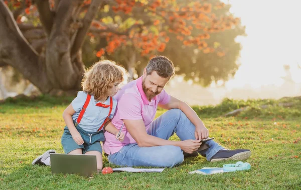 Son with father learning outdoor by studying online and working on laptop in green field. Childhood and parenthood kids concept. — Stock Photo, Image