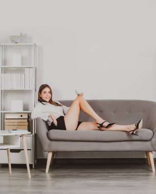 Seksi bacaklı baştan çıkarıcı sekreter ofiste kanepede oturur. Kahve içen seksi iş kadını. Şehvetli kız çalışan..