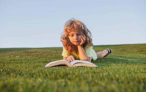 Söt pojke läser bok utomhus på grönt gräs fält. Smart barnbok, ligger på gräs sommar park på himlen bakgrund. Porträtt av smarta barn. — Stockfoto