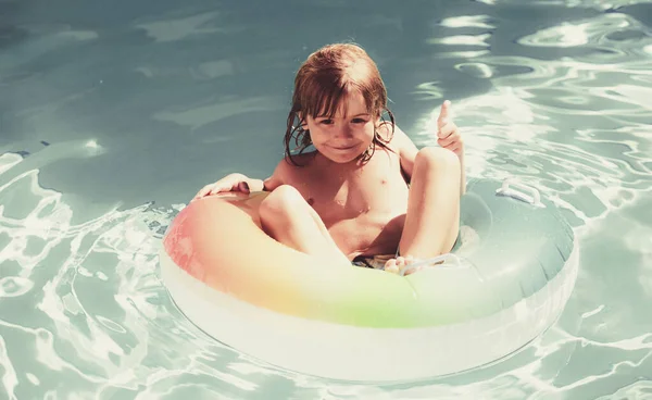 Bonito menino criança engraçada com polegar para cima em um terno de natação colorido e óculos de sol relaxante. Criança com anel de brinquedo flutuando em uma piscina se divertindo durante as férias de verão em um resort tropical. — Fotografia de Stock