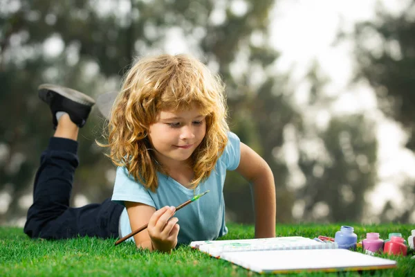 Ребенок рисует в парке лежа в траве весело на фоне природы. Детское хобби, счастливое детство. — стоковое фото