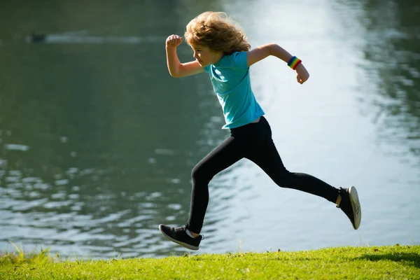 Barnpojken springer eller joggar nära sjön på gräs i parken. Sportig pojklöpare som springer i sommarparken. Aktiva barn, sportbarn. Jogging hjälper kroppen att vara stark. — Stockfoto