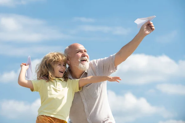 Neto jovem e avô velho com avião de papel de brinquedo contra o fundo do céu de verão. Menino com sonhos de voar. — Fotografia de Stock