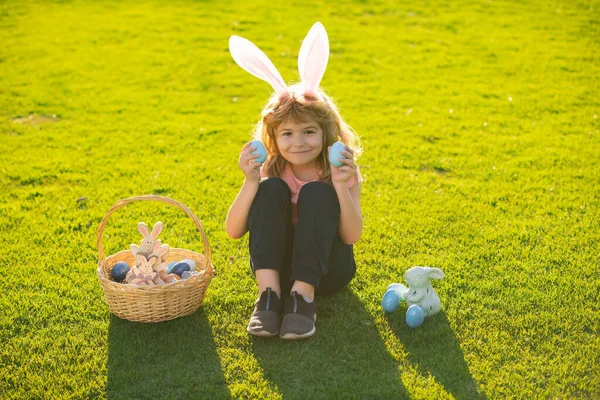 Chłopiec w stroju królika z króliczymi uszami polujący na wielkanocne jaja na trawie w parku wiosennym. Zabawny chłopiec, króliczki wielkanocne. — Zdjęcie stockowe