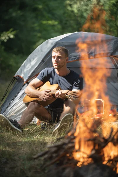 Homem romântico acampando ao ar livre e sentado perto da tenda. Homem bonito tocando guitarra na floresta com fogueira. Música country. Um tipo perto de um incêndio na floresta, fim-de-semana perto da fogueira. — Fotografia de Stock