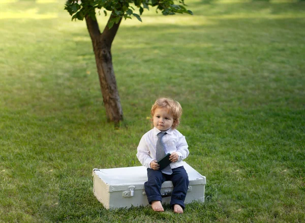 Baby kind in pak en stropdas zitten op koffer de zomer op de natuur in het park buiten. Leuke baby met koffer buiten. Concept reis met kinderen. Kleine reiziger. — Stockfoto
