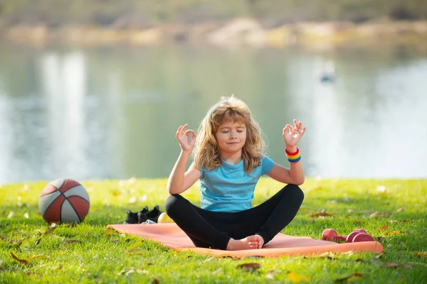 Niño pequeño sentado en la alfombrilla practicando yoga de meditación en el parque al aire libre. Niño practicando yoga pose, loto pose yoga. — Foto de Stock