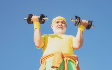 Mavi arka planda izole edilmiş dambıllı kıdemli fitness adamı eğitimi. Spor salonundaki son sınıf öğrencisi ağırlık çalışıyor. Kıdemli adam ağırlık kaldırıyor..