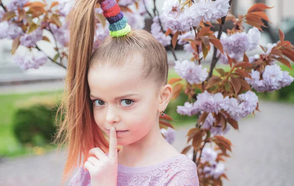 Positief schoolmeisje met een lang kapsel die haar vinger naar de lippen houdt. Kindergeheimen. Bloeiende sakura kers. — Stockfoto