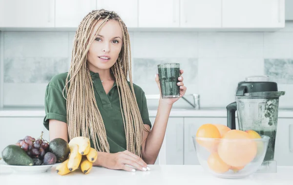 La mujer positiva sostiene la taza de batido verde fresco. Eco estilo de vida. Mujer joven bebiendo batido verde en la cocina con frutas y verduras. Menú de comida vegana y desintoxicación. — Foto de Stock