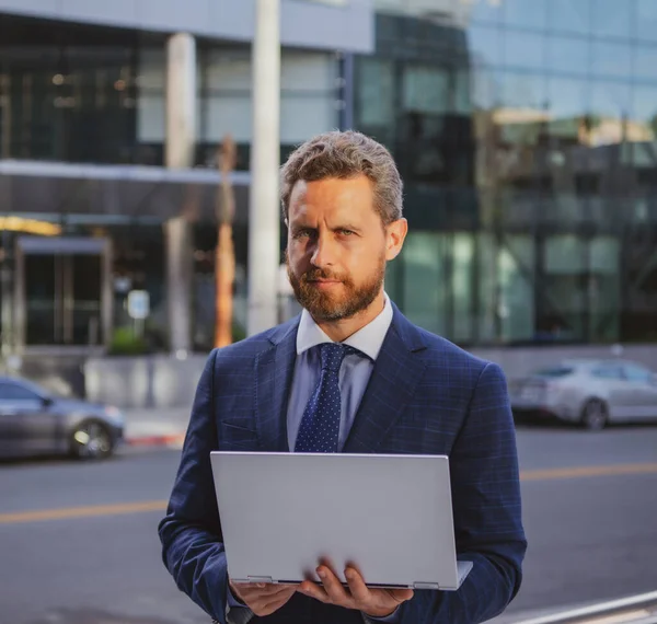 Geschäftsmann mit Notebook im Freien. Selbstbewusster Wirtschaftsexperte. Schöner Mann im Anzug hält Laptop vor Bürohintergrund. — Stockfoto