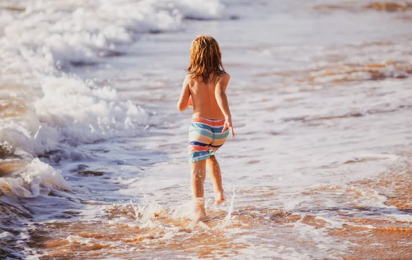 Vista posteriore del ragazzo che corre sulla spiaggia vicino al mare. Eccitato bambino stupito divertirsi con la corsa attraverso l'acqua in mare o mare. — Foto Stock