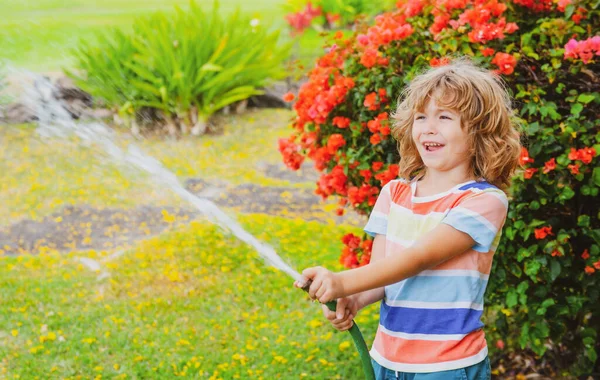 Netter kleiner Junge, der an Sommertagen Blumen im Garten gießt. Kind mit Gartenschlauch. Lustiges Kind gießt Pflanzen im Hofgarten. — Stockfoto