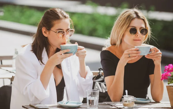 Two beautiful girls with cups coffee in summer cafe. Girl friends in cafe outdoor. Outdoors portrait of two young beautiful women friends drinking coffee. — Foto Stock