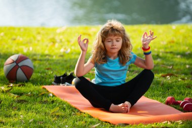 Çocuk yoga yapıyor, spor kıyafetleri giyiyor, dışarıda tişört giyiyor. Yoga sırasında meditasyon yapan çocuklar. Küçük çocuk nilüfer çiçeği pozunda meditasyon yapıyor. Jimnastik, çocuklar için meditasyon.