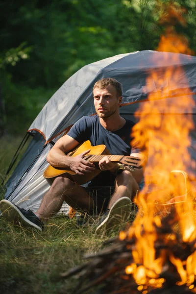 Jovem sentado perto da tenda turística e tocando guitarra. Tempo romântico em um acampamento de montanha. Turismo e caminhada, estilo de vida de viagem. Bonito modelo atraente homem tocando guitarra na floresta com fogueira. — Fotografia de Stock