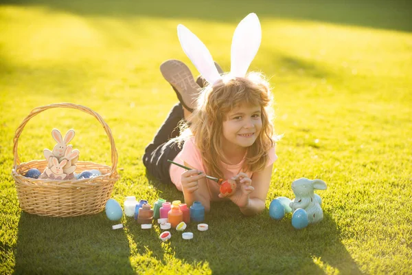 Дети празднуют Пасху. Мальчик в костюме кролика с кроличьими ушами, рисующий яйца на открытом воздухе. — стоковое фото