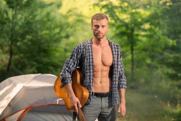 Música al aire libre. Hombre sexy con guitarra en camisa a cuadros, camping hipster y senderismo. Tipo romántico acampando al aire libre cerca de la tienda. — Foto de Stock