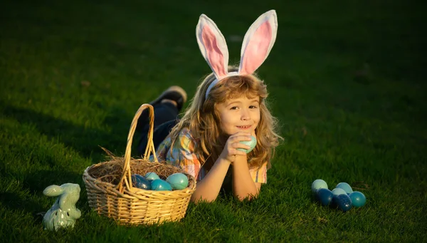 Enfant chassant des œufs de Pâques dans la cour, pondant sur l'herbe verte. Lapin enfants avec lapin oreilles de lapin. — Photo