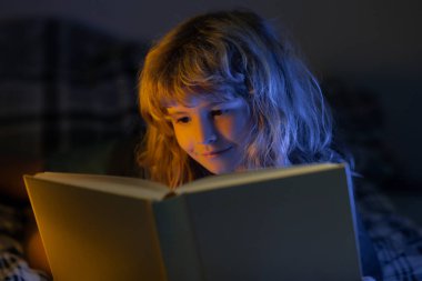 Çocuk karanlık odada masal okuyor. Kitap okuyan şirin bir çocuk. Açık kitabı olan bir çocuk. Ev okulu dersi. Evde kitap okuyan konsantre küçük çocuk..