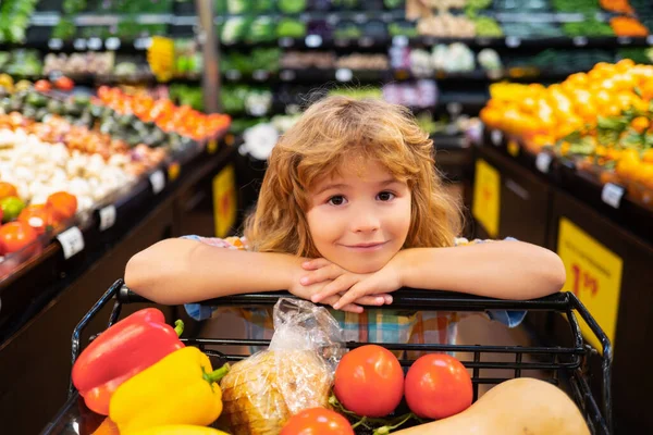 Piccolo ragazzo carino con carrello pieno di verdure fresche biologiche e frutta in negozio di alimentari o supermercato. Cibo sano per bambini. Bambino nel supermercato compra verdure. — Foto Stock
