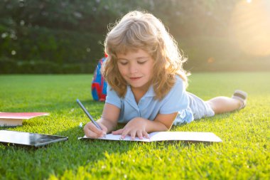 Not defterine kalem yazıp çimlere uzanan sevimli çocuk. Çocuk yaz parkında kitap okuyor..