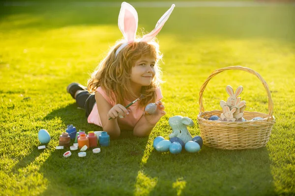 Niño con traje de conejo con orejas de conejo cazando huevos de Pascua en la hierba en el parque de primavera. Los niños de Pascua con orejas de conejo y sobre el fondo de hierba. — Foto de Stock