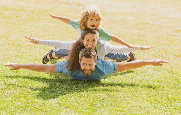 Family lying on grass in park. Fly concept, little boy is sitting pickaback while imitating the flight. — Stock Photo, Image
