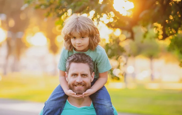 Padre llevando a su hijo de vuelta al parque. Retrato de un padre feliz dando a su hijo un paseo a cuestas sobre hombros. Lindo niño con papá al aire libre. — Foto de Stock