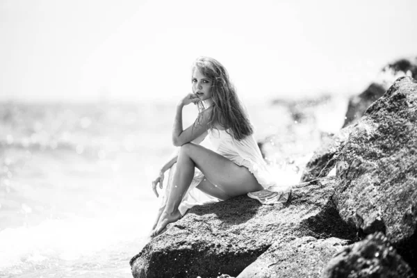 Sensuell kvinna sitter på sandstenig strand mot blå vågor. Sommarflickans dröm. Attraktiv ung kvinna bär sexig sommarklänning på en strand. — Stockfoto